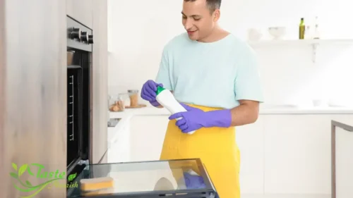 "Hand wearing yellow glove applying oven glass cleaner paste with blue sponge to dirty oven door interior"
